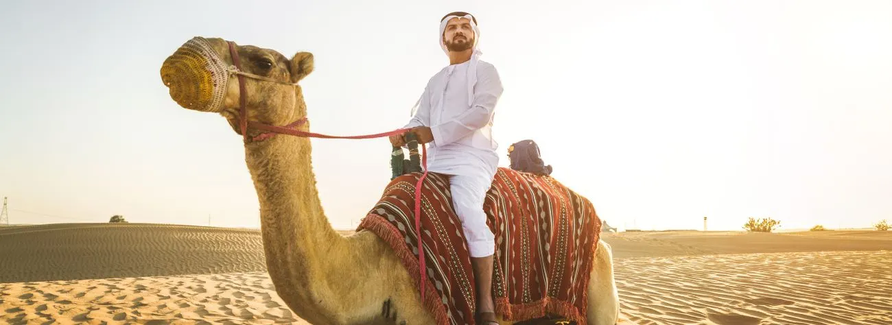 Man riding a camel in the desert at sunset near LuxuryAirs Camp, wearing traditional Arab attire