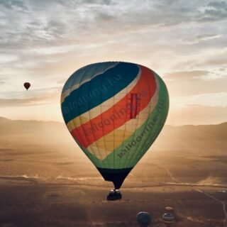 A colorful hot air balloon floating over a vast desert landscape during a golden sunrise, with distant mountains and a soft glow on the horizon.