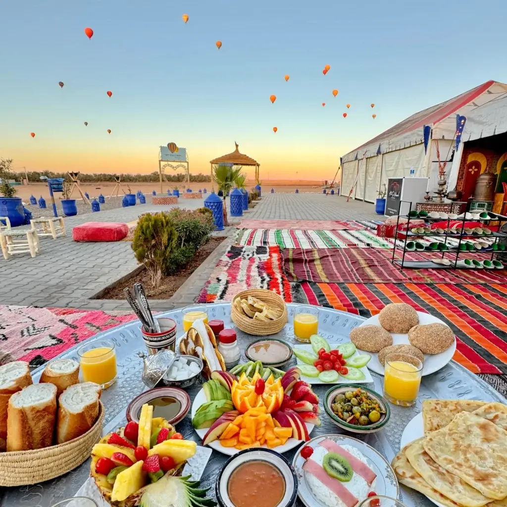 Traditional Moroccan breakfast spread with fresh fruit, pastries, and orange juice at a desert camp with hot air balloons in the sky – LuxuryAirs