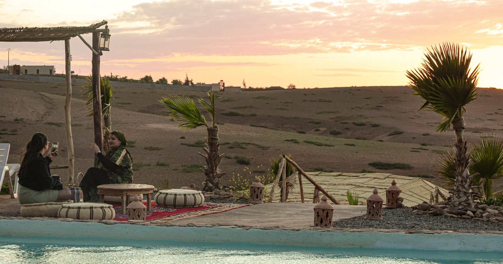 Two women sit together under a warm sunset sky in the Agafay Oasis, with a serene pool beside them and desert hills in the background.