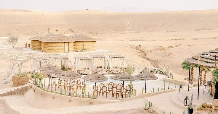 Luxury desert resort with eco-style straw huts, poolside lounge area, and scenic dunes in Morocco or Middle Eastern landscape.